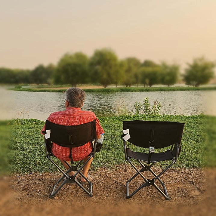 Person sitting in a folding chair by a tranquil lake with an empty chair beside them.