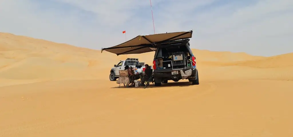 Two off-road vehicles are parked in a desert with an awning providing shade. People sit underneath on folding chairs in the sandy landscape, surrounded by rolling dunes under a partly cloudy sky. A small flag is attached to one vehicle.