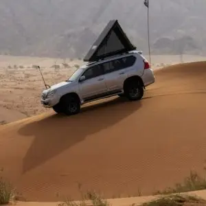 A silver SUV fitted with a Chicari Roof Top Tent old ascends a steep sand dune in the desert. The overcast sky looms above, with mountains visible in the distance.