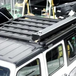 A white SUV with a black roof rack features the Road Shower and an awning on top, parked indoors. Other vehicles and a green area with chairs can be seen in the background.