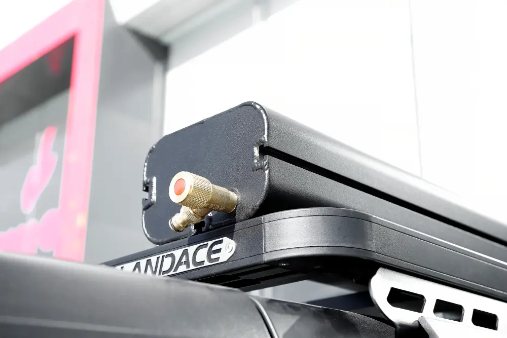 MG_2778.jpg A black rectangular rooftop cargo box called the Road Shower is seen mounted on a vehicle. It has a metallic valve with a red center at the front, and part of the vehicles roof rack is visible underneath.