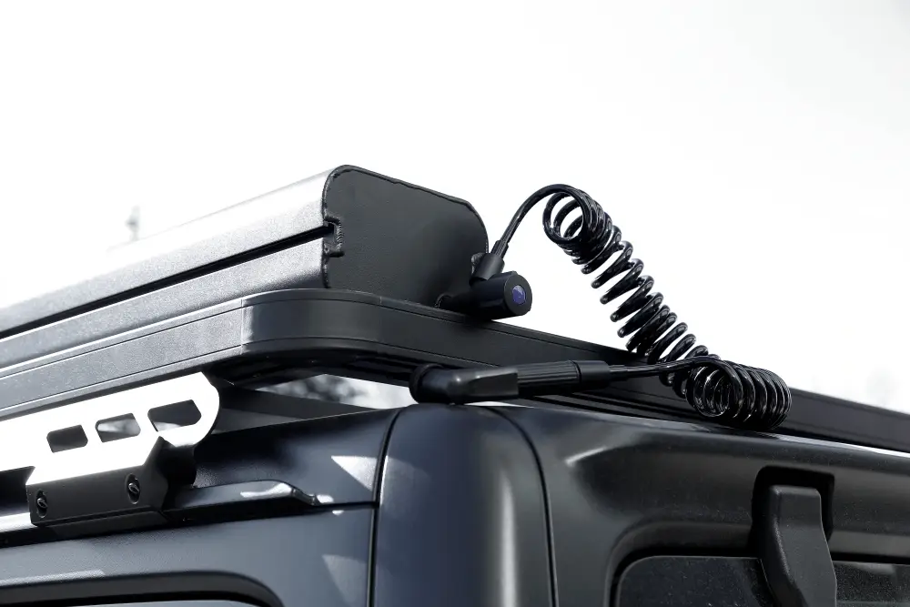 MG_2780.jpg Close-up of a vehicle roof rack with a mounted Road Shower and coiled cable attached. The image highlights the connection point and cable details against a blurred background.