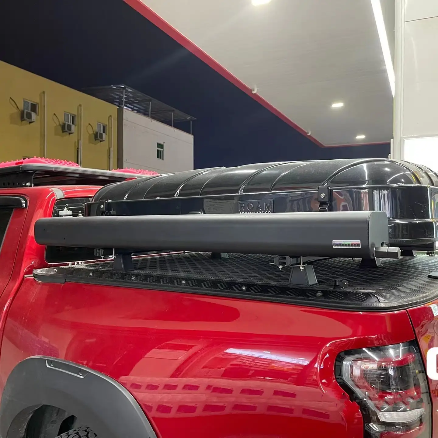 微信图片_202205091401581.jpg A red pickup truck with a black cargo carrier and roof rack featuring a Road Shower is parked at a well-lit gas station at night, with exterior units visible on the building in the background.