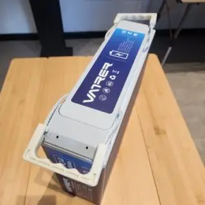A blue and white 100Ah Slim Lifepo4 Battery labeled VANBREE, featuring side handles, sits on a wooden surface in a well-lit room near a metal table. Various icons are visible on the label.