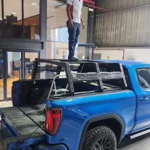 A man stands on a Chicari Bed Rack mounted in the bed of a blue pickup truck inside a garage or workshop with open doors and industrial lighting.
