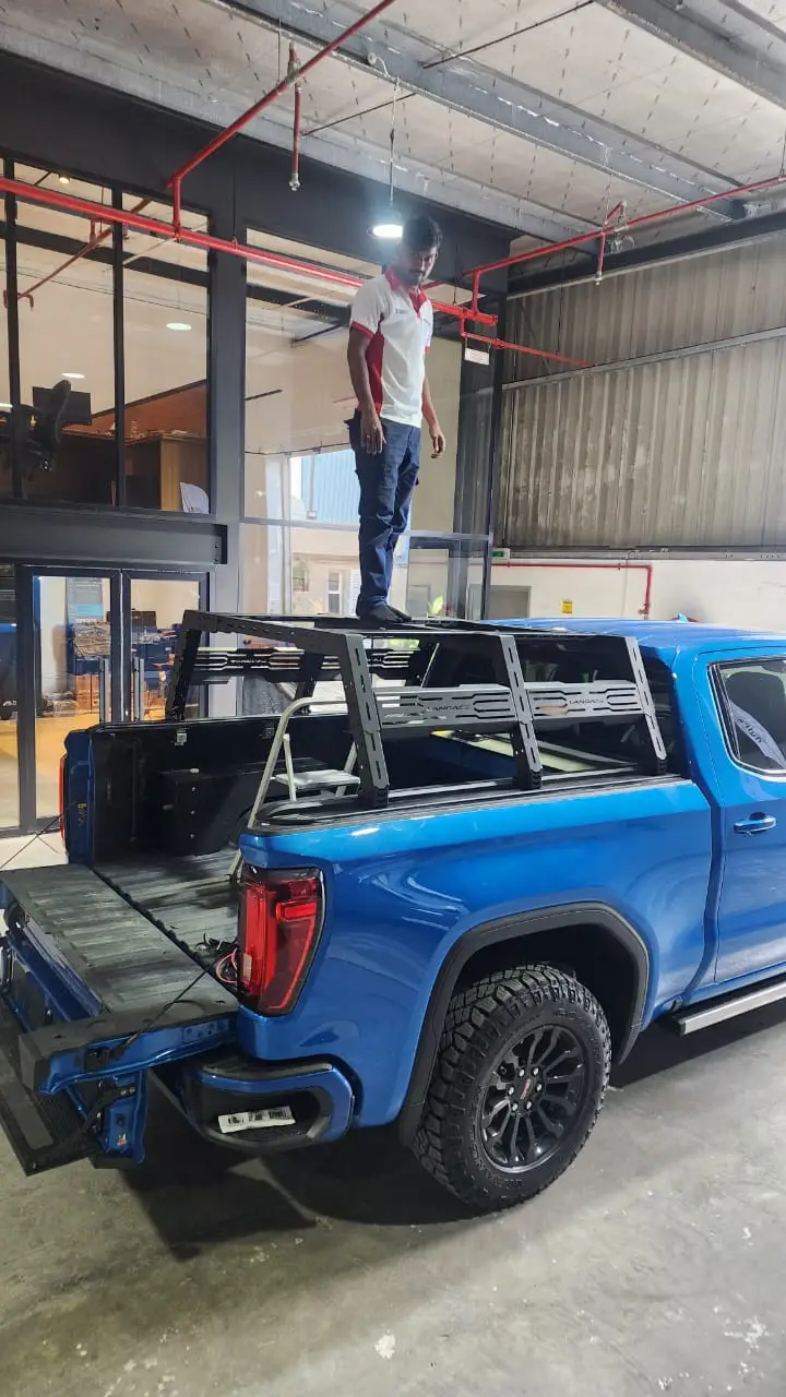 bedrack_gmc A man stands on a Chicari Bed Rack mounted in the bed of a blue pickup truck inside a garage or workshop with open doors and industrial lighting.