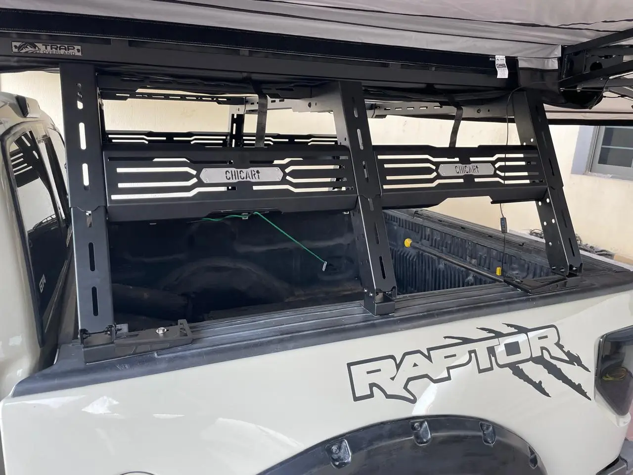 bedrack_raptor A close-up shows a white pickup with a Raptor decal, featuring a Chicari Bed Rack in black metal installed over the empty truck bed and an extended awning above it.
