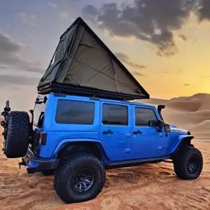 A blue off-road vehicle with a rooftop tent is parked on sandy dunes at sunset, with a dramatic sky and hills of sand in the background.