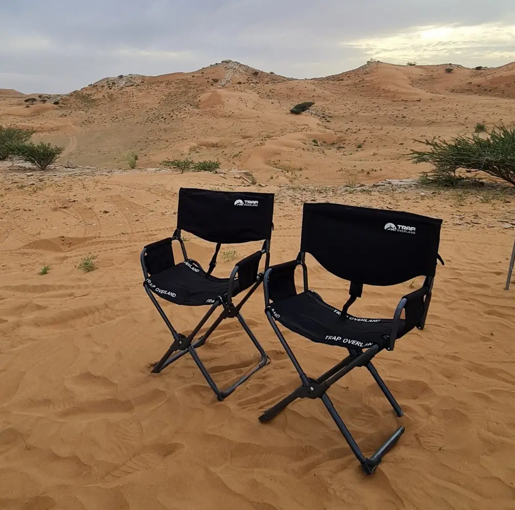 Two black folding chairs are set up on sandy desert ground, with dry shrubs and rolling dunes in the background under a cloudy sky.