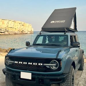 A blue Ford Bronco with a rooftop tent parked near a rocky shoreline, with calm sea water and a large cliff in the background under clear skies.