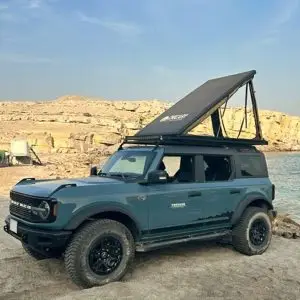 A blue off-road SUV with a raised rooftop tent parked on sandy ground near rocky cliffs and a body of water under a clear sky.