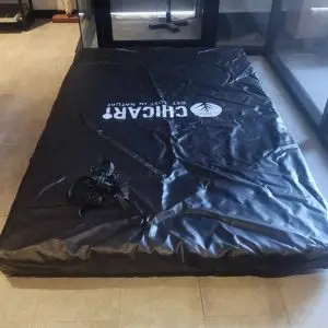 A large black Chicari RTT Cover with a logo, closed on a tiled indoor floor by a glass wall. “CHIGARU” is printed on it and straps are visible.