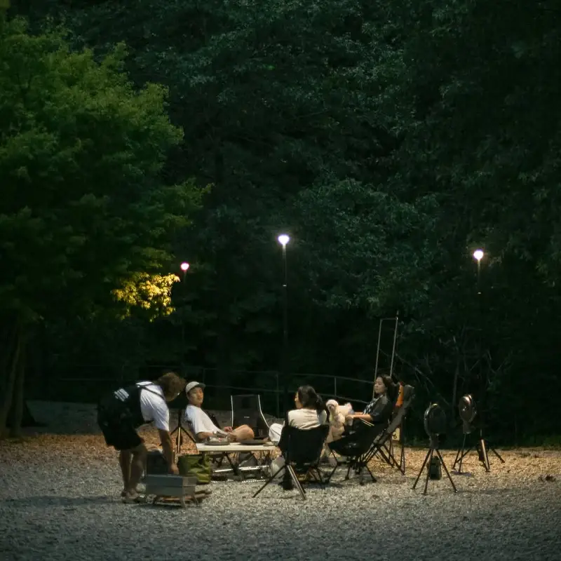 finel_camping_VIP_._._._N7_8 Four people sit around a campsite at night, surrounded by trees and illuminated by the Finel N7 Camping Light for a romantic ambiance. One person stands nearby, setting something down as the calm, relaxed scene unfolds.