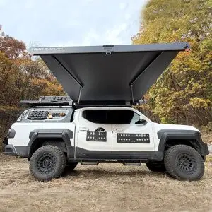 A white off-road vehicle with large rugged tires and roof cargo gear is equipped with the Fast Awning, extended from its roof. It’s parked outdoors in the UAE, with autumn trees displaying colorful leaves in the background.