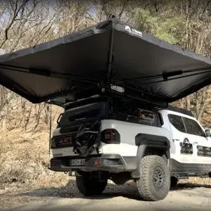 A white off-road truck parked outdoors with a large, black, 270-degree awning extended from its roof rack, surrounded by dry trees and scattered leaves.